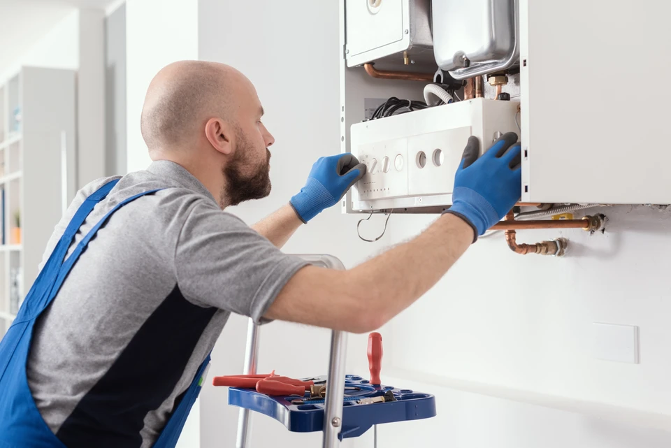 Technicien en combinaison bleue et gants de protection réparant une chaudière murale domestique dans un intérieur résidentiel