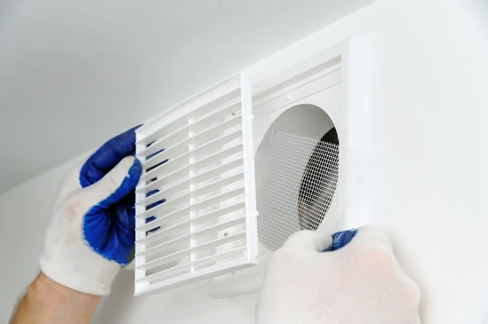 Intervention de maintenance dans un logement, technicien portant des gants de protection bleus et blancs proc&eacute;dant au d&eacute;montage d'une grille de ventilation murale blanche