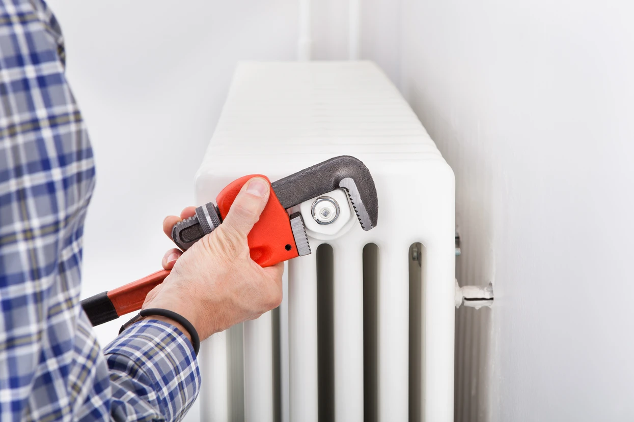Un plombier en chemise à carreaux bleus utilise une clé à molette rouge pour serrer un raccord sur un radiateur blanc