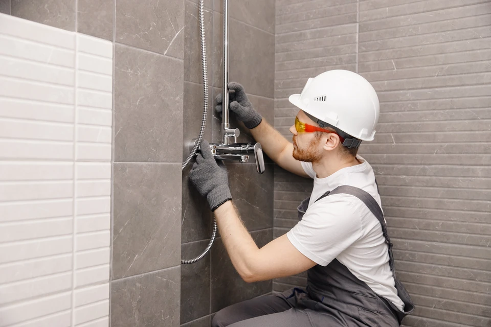 Plombier professionnel en casque blanc et lunettes de protection installe une colonne de douche thermostatique dans salle de bain carrelée