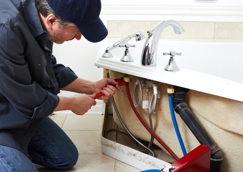 Plombier professionnel travaillant sous un lavabo de salle de bain, utilisant une clé à molette rouge pour réparer ou installer la robinetterie