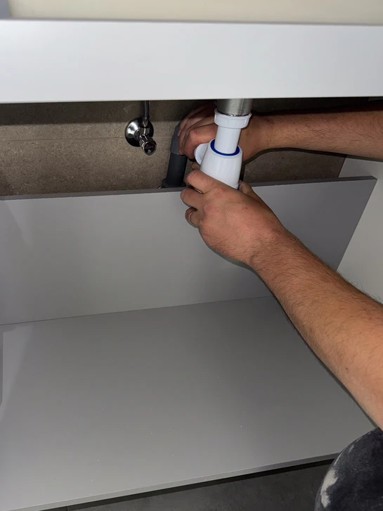 Photographie d'un plombier-chauffagiste dans une salle de bains en cours de r&eacute;novation, expliquant les d&eacute;tails du projet &agrave; un client. L'artisan pointe des &eacute;l&eacute;ments du chantier (canalisation, installation de douche, &eacute;quipements sanitaires) tandis que le client l'&eacute;coute attentivement. L'environnement montre un chantier organis&eacute; et contr&ocirc;l&eacute;, avec des outils professionnels visibles. La sc&egrave;ne doit transmettre la confiance, la clart&eacute; des explications et la ma&icirc;trise professionnelle du projet.